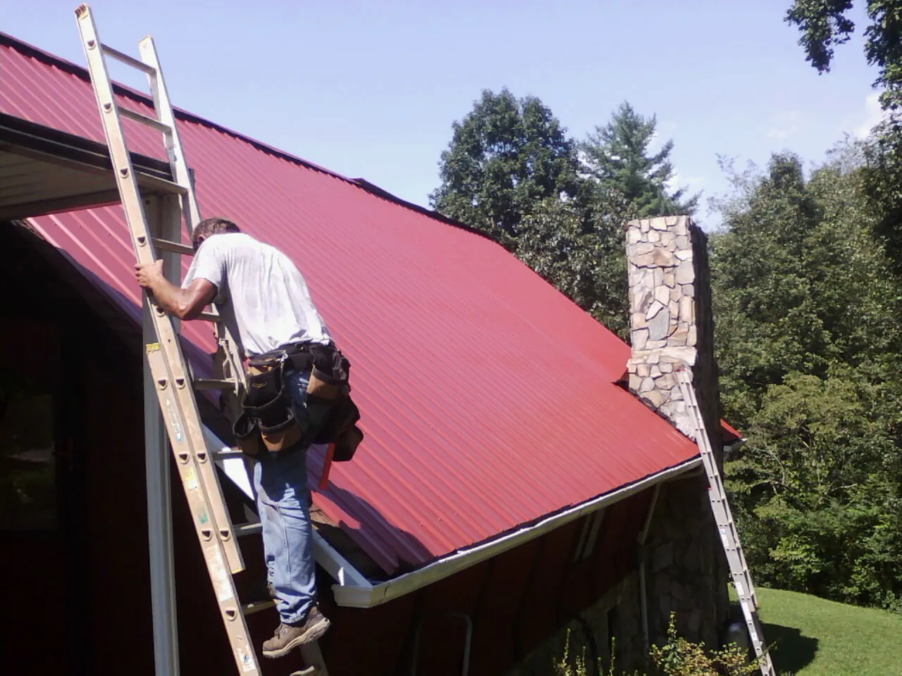 Roofer on a ladder beside a red metal roof and stone chimney — Soffit & Fascia Repair by Roofing Brothers in Wolfeboro